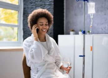A person on the phone smiling while seated near an IV pole in a room with large windows