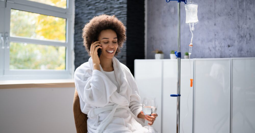 A person on the phone smiling while seated near an IV pole in a room with large windows