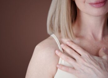 Woman with short blonde hair lightly touching her chest area against a brown background