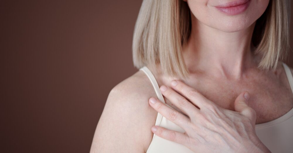 Woman with short blonde hair lightly touching her chest area against a brown background