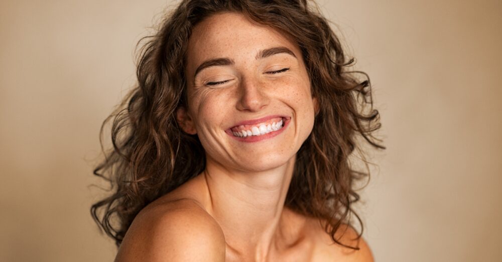 Woman with freckles smiling with her eyes closed against a neutral background
